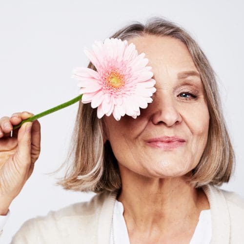 Cheerful elegant elderly woman holding a flower near the face on a gray background